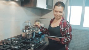 woman cooks soup in the kitchen prevents the water in the pot and looks at the recipe on the digital tablet. woman in the kitchen slow motion video. girl mom in the kitchen indoors preparing food