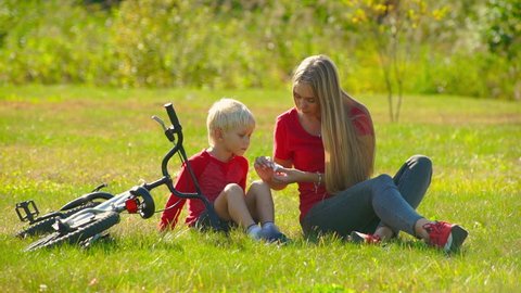 Slow motion shot of a young woman applying cut plaster on a knee of her little son and a bicycle laying next to them