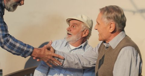 Close up of the two old Caucasian retired men neighbors talking while sitting and resting on the bench on a sun their friend coming to them and shaking hands. Outdoors.
