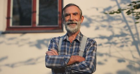 Portrait shot of the senior Caucasian man with grey hair, beard and mustache crossing his hands in front and looking straight to the camera. Outdoors.