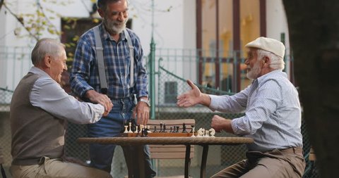 Caucasian senior men neighbors having a chess game in the yard and their old friend coming to join them, greeting, shaking hands and sitting next to them. Outdoors.