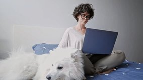 Smiling brunette woman using laptop computer while sitting on bed with her little dog at home
