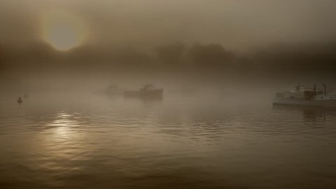 A lobster fishing boat moves quietly through the water and early morning light at sunrise in a foggy and quaint New England harbor.