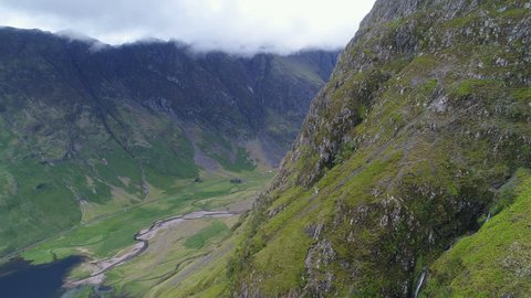 Drone shot flying past steep mountain cliffs reveals road cutting through spectacular nature valley in Glencoe, Scotland