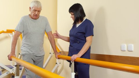 Tilt down shot of elderly man walking between handrails and supporting his weight while learning to walk with help of female physiotherapist