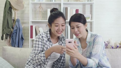 slow action of relaxed friends using a generic mobile phone together sitting on a sofa in the living room at home. girls watching funny video movie laughing cheerfully in cozy apartment.