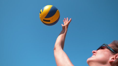 SLOW MOTION, CLOSE UP, DOF: Beautiful young Caucasian woman playing volleyball strikes the ball above her head on a sunny day. Girl wearing blue sunglasses serves the ball during beach volleyball game