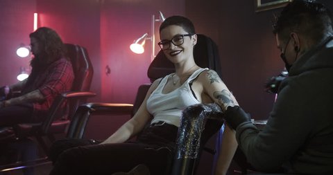 Young people in tattoo studio. Slow Motion portrait of a pretty short hair teen girl getting a tattoo in arm at beauty parlour. Young gay woman gets tattooed. Portrait of woman looking at camera.