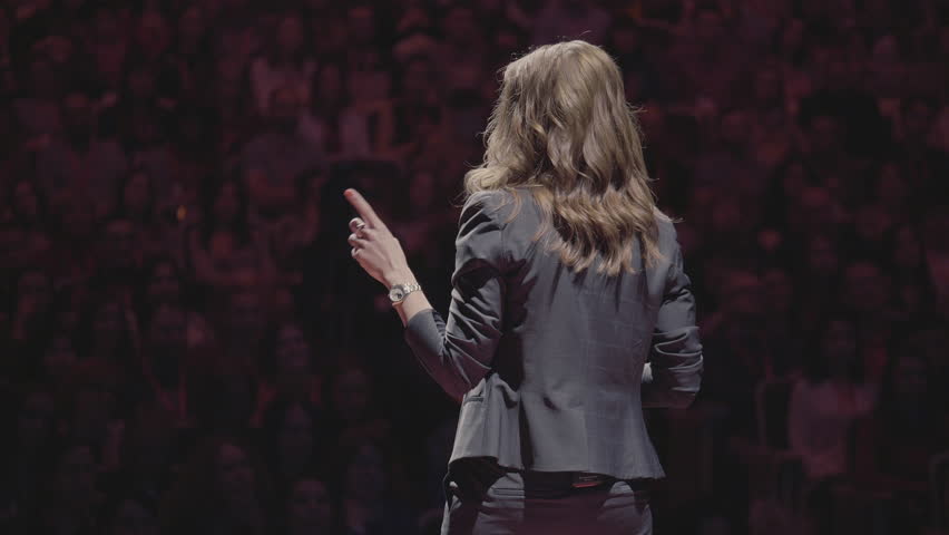 The female financial coach emotional gesturing talks from the stage with spectators at forum. Too many anonymous persons workers and students seat in large auditorium and watch workshop background | Shutterstock HD Video #1020705976