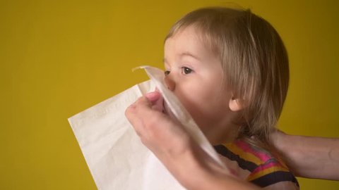 Mother helping little girl to blow nose by napkin indoor, view in profile. Woman keeping napkin near her daughter face while toddler blowing her nose. Adult helps baby taking care during sickness