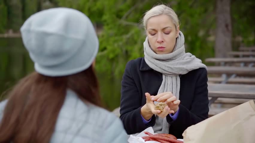 Portrait of a beautiful blonde woman wearing black coat and gray scarf eating tasty sausages with a friends outdoor in park cafe.