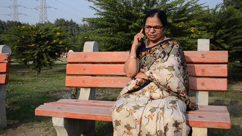 Pan shot of Senior Indian woman with glassses wearing traditional saaree laughing and speaking on phone, sitting in a park in Delhi. Concept - Digital literacy for senior citizens of India