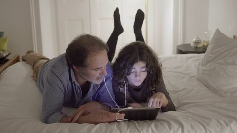 Father and daughter lying in bed, using tablet computer. Each of them wearing earphone, they communicating with each other, reaching conclusion. Father explaining something. Technology concept.