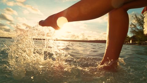 SLOW MOTION, CLOSE UP, LENS FLARE: Unrecognizable playful woman having fun kicking the refreshing ocean water while sitting on the pier on a sunny summer evening in Cook Islands. Girl splashing water.