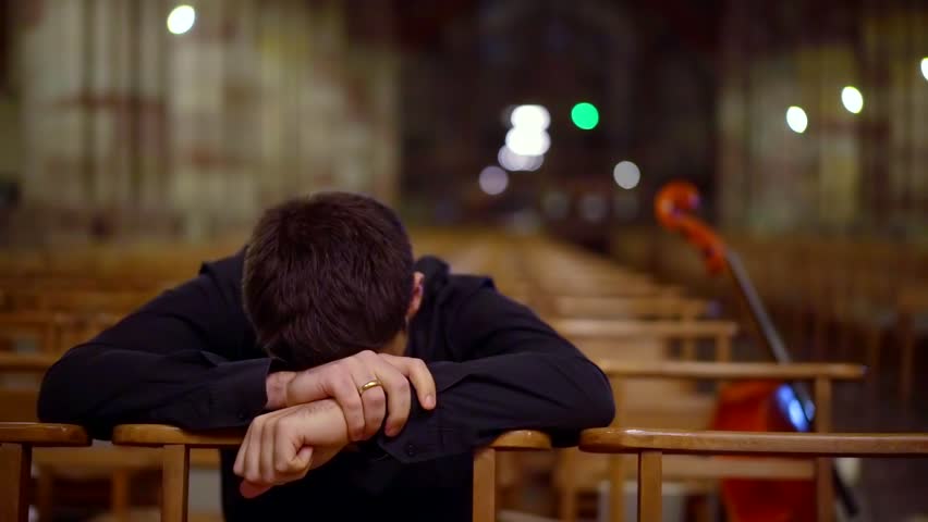 sad man is sitting bowing head on crossed hands on chair in empty hall of church, thinking