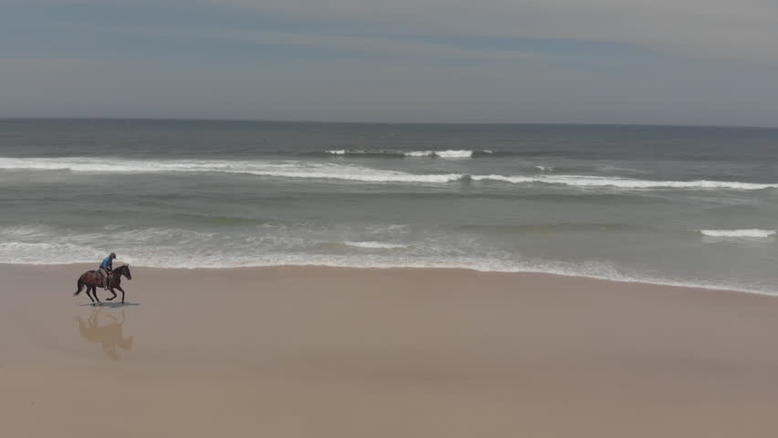 Side on follow shot of male horse rider on sandy beach with waves lapping in to coast.