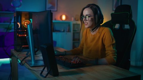 Excited Gamer Girl in Headset with a Mic Playing and Winning in Online Strategy Video Game on Her Personal Computer. Room and PC have Colorful Warm Neon Led Lights. Cozy Evening at Home.