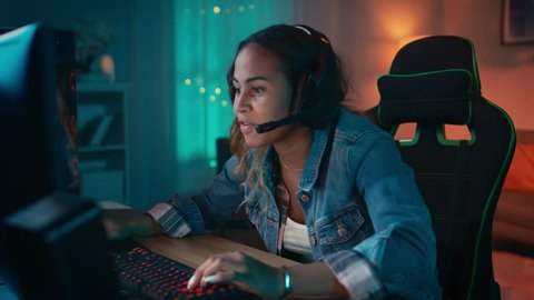 Excited Gamer Girl in Headset with a Mic Playing Online Video Game on Her Personal Computer. She Talks to Other Players. Room and PC have Colorful Warm Neon Led Lights. Cozy Evening at Home.