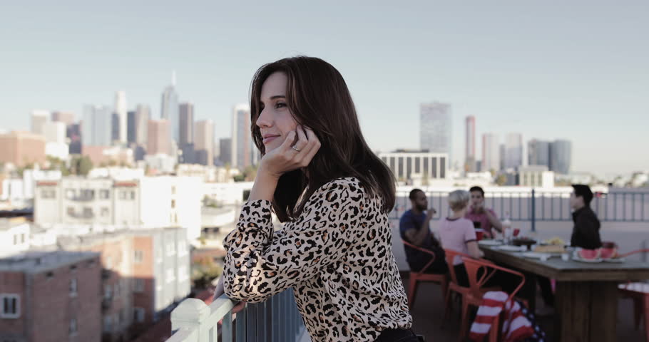 Young adult female looking out across city at a rooftop party and then smiling to camera