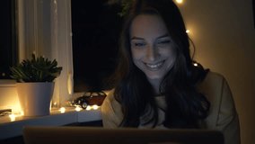 Young beautiful woman smiling while using laptop computer, watching movies and videos while sitting near the window at night. Internet, social network. Close up
