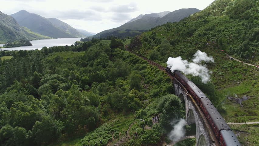 Beautiful drone flight tracking historic steam train near the famous Glenfinnan viaduct, among beautiful green mountain landscape in Scotland