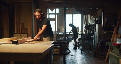 Carpenter woodworker cuts wood on saw in factory atelier during the day. Long to medium shot with warm lighting on 4K RED camera.