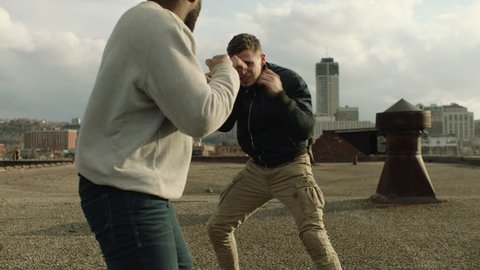 Buff fighter man punches and knocks out another man on a warehouse rooftop in overcast sunlight. Medium shot in 4K with an Alexa Mini camera