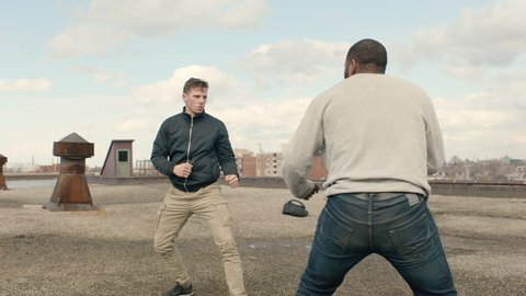 Caucasian man and African American man fight on a warehouse rooftop with hard kicks and punches in overcast sunlight. Medium shot in 4K with an Alexa Mini camera