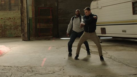 Two men fight over possession of knapsack using martial art techniques, in front of an abandoned camper trailer in moody lighting. Medium shot in 4K with an Alexa Mini camera