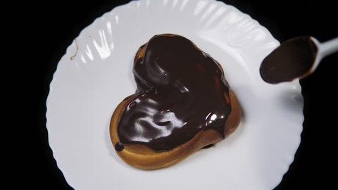 Someone decorates dark topping white plate with a bun in the shape of a heart covered with chocolate.