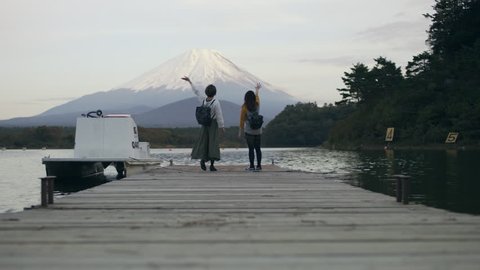 Wide shot on 4k RED camera. Excited Japanese women running up a dock on the water to see Mount Fuji and happily waving at it with soft natural lighting. Stock Video