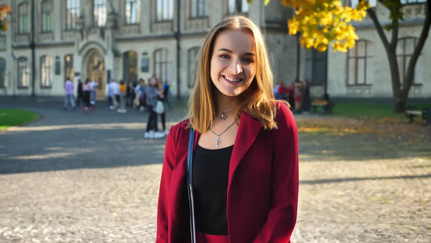 Young female student laughs, looking on a camera standing on the background of university