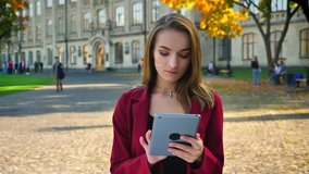 Young attractive female, student browsing on the tablet and showing screen to the camera, thumbs up, smiling, outdoors. Outside the university