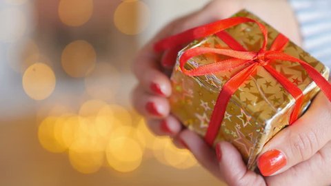 holidays, close-up of a girl's hand carefully hold a small box with a gift wrapped in golden paper with a red ribbon, on a bright festive background