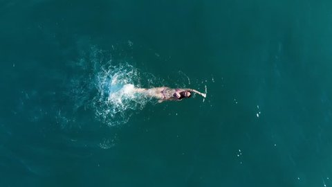 Young woman swim at sea, lie on back and using only legs, white splashes from quick movement. Top-down aerial shot, camera move up and then begin to spin around, reveal wide view