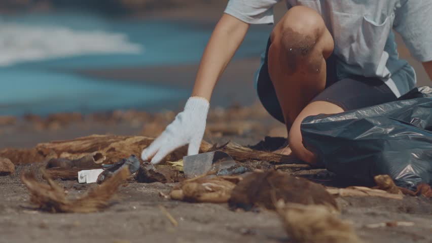 Close up of volunteers sit and picking up garbage on the beach. Cleaner collecting garbage on the black sand beach into black plastic bag. Volunteers cleaning the beach. Tidying up rubbish on beach Royalty-Free Stock Footage #1020794275