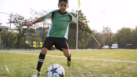 Young boy in the green uniform making exercise with soccer ball on the football field on the sunshine background. Dolly shot.