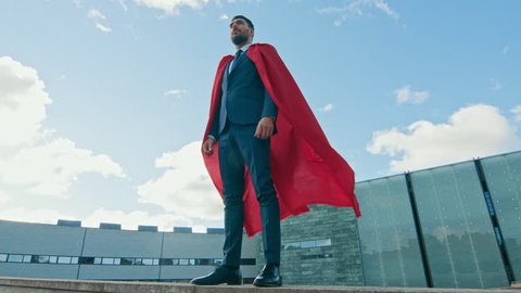 Superhero Businessman With Red Cape Blowing in the Wind Stands on the Roof of a Skyscraper Ready to Make Business Transactions and Save the Day. Low Angle Shot.