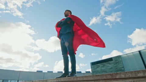 Superhero With Red Cape Blowing in the Wind Stands on the Roof of a Skyscraper, Crossed Arms, Ready to Make Business Transactions and Save the Day. Low Angle Shot.