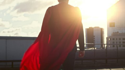 Superman WIth Red Cape Blowing in the Wind Walks on the Roof of a Skyscraper, Looking into the Sunset, Ready to Save the Day. Following Back View Slow Motion Shot.