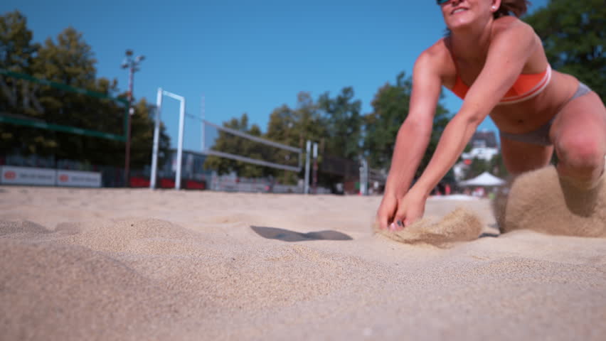 SLOW MOTION, CLOSE UP, DOF: Smiling Caucasian female volleyball player dives and strikes ball with her hands. Young woman on active summer vacation playing beach volleyball on a sunny day. Sand flying