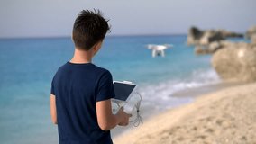 Young boy operating a flying drone on a coast of a sea. An early teen male child, wearing a deep blue T-shirt seen from his back, is holding a remote control unit in his hands and manipulating a white