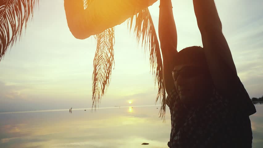 Young happy man in sunglasses hangs on a palm tree at amazing sunset on the beach. slow motion. 1920x1080