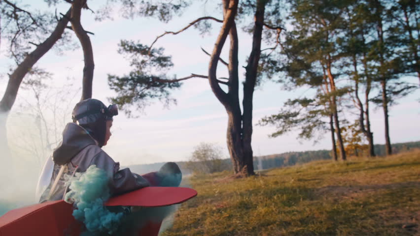 Camera follows little happy aviator boy running in forest wearing cardboard plane costume with color smoke slow motion.