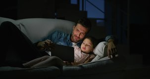 Portrait of happy father and daughter using a tablet on sofa in the evening in slow motion. Shot with RED camera in 8K. Concept of family entertainment, education, technology