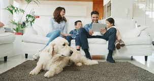 Portrait of happy family with a dog having fun using tablet on sofa in living room in slow motion. Shot with RED camera in 8K. Concept of  family entertainment, education, technology, love for animals