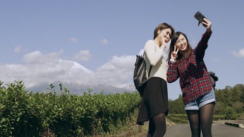 Wide shot of two happy Japanese friends taking a picture together with a mobile phone with Mount Fuji in the background in a tea plantation with soft natural lighting.  Stockvideó