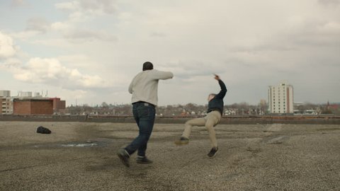 Two men fight and get knocked down and get up on rooftop with skilled kicks and punches in overcast sunlight. Medium shot in 4K with an Alexa Mini camera