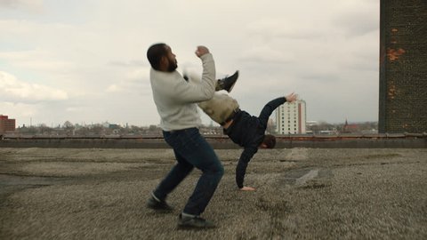 White man is knocked to the floor as he fights an African American man on a warehouse rooftop with skilled kicks and punches in overcast sunlight. Medium shot in 4K with an Alexa Mini camera
