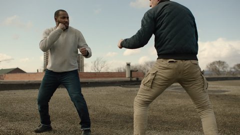 Angry man is knocked to the floor as he fights a bigger man on a warehouse rooftop with skilled kicks and punches in overcast sunlight. Medium shot in 4K with an Alexa Mini camera